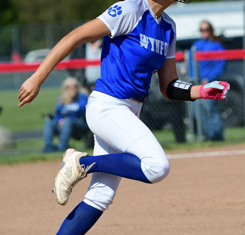 Female softball player in blue and white uniform running on a dirt field, wearing helmet and pink glove on her right hand.