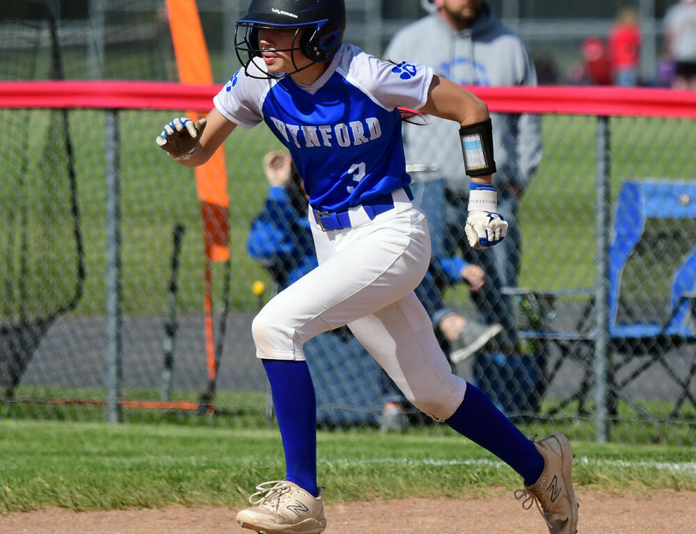 Softball player in a blue Winford uniform sprinting along the dirt baseline with a helmet on and gloves ready at the field edge.