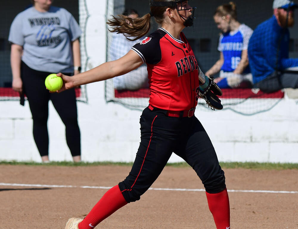 Softball pitcher in a red and black uniform winds up to throw a pitch on the dirt mound, holding a bright yellow ball.