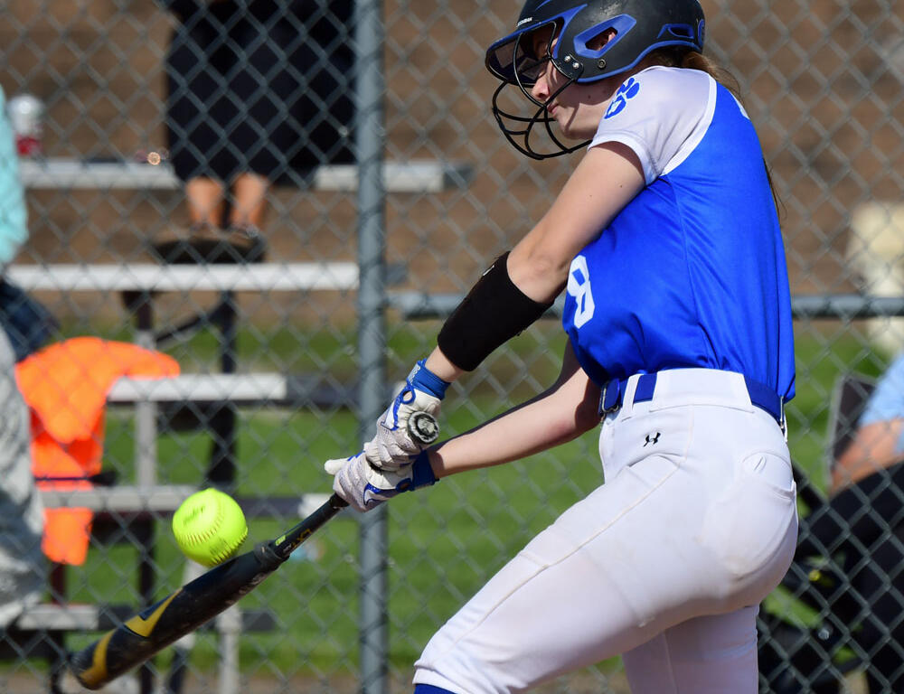 Female softball player in a blue jersey and white pants swings a bat at a bright yellow ball near a chain-link fence.