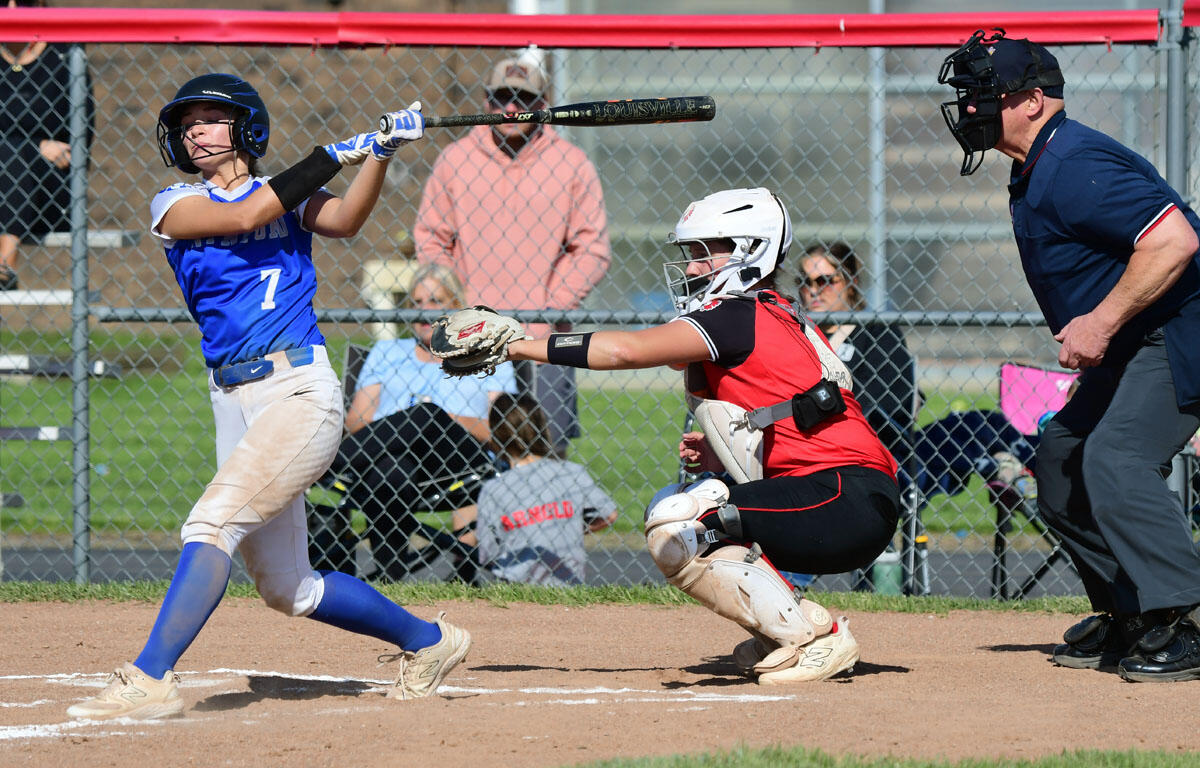 Female softball batter in a blue uniform (#7) swings at a pitch as the catcher in red gear crouches behind home plate and an umpire watches nearby.