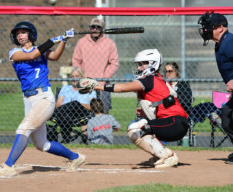 Female softball batter in a blue uniform (#7) swings at a pitch as the catcher in red gear crouches behind home plate and an umpire watches nearby.