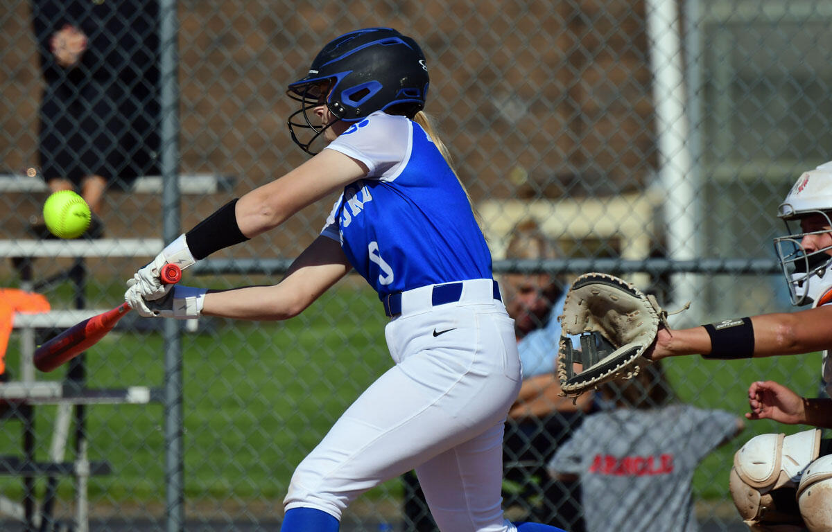 Female softball player in blue and white swings a red bat at a pitched ball near the catcher and fence in background.
