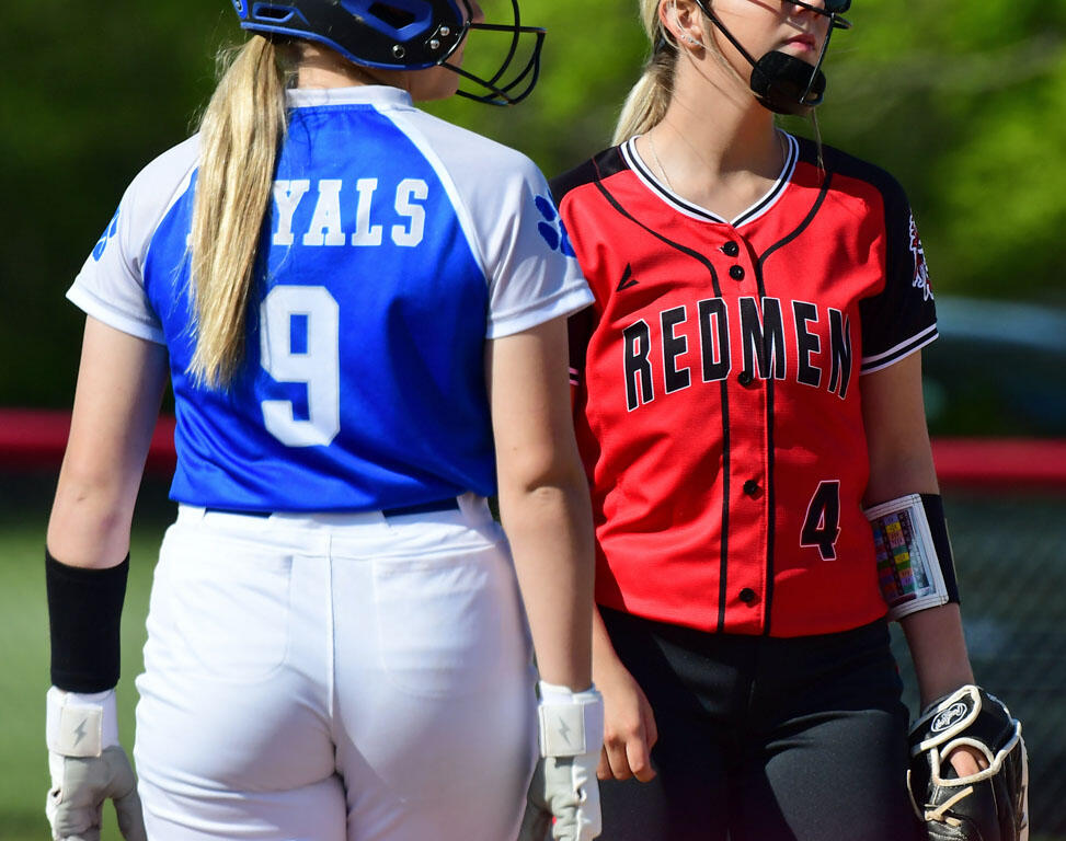 Two female softball players on a field facing each other: blue Royals jersey number 9 with helmet, white pants, and gloves, beside a red Redmen jersey number 4 with mask and glove.