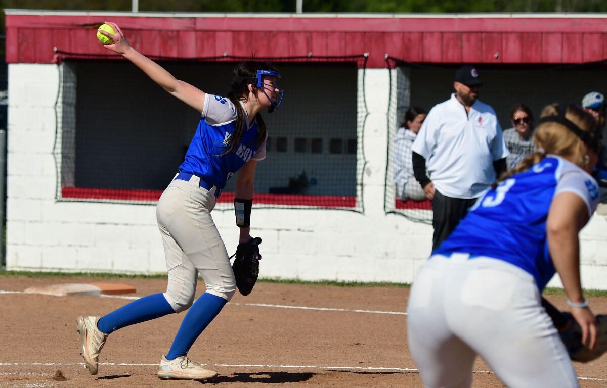 Softball pitcher in a blue jersey throws from the mound with teammates in the background.