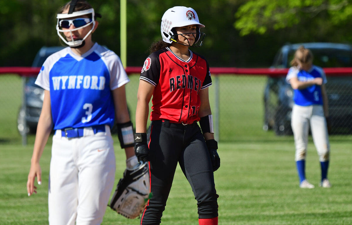 Center: a female softball player in a red Redmen jersey and helmet stands ready on a sunny field with black pants and red socks.