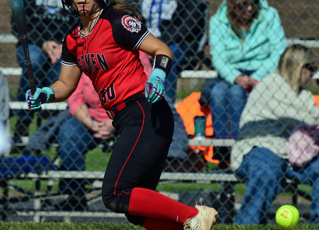 Female softball player in a red-and-black uniform swings a bat during a game, helmet on, spectators behind a chain-link fence.