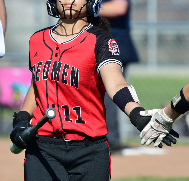 Young female softball player in a red and black uniform (#11) standing on the field, holding a bat and wearing a white helmet with a mascot logo, ready to run or greet teammates after a play.