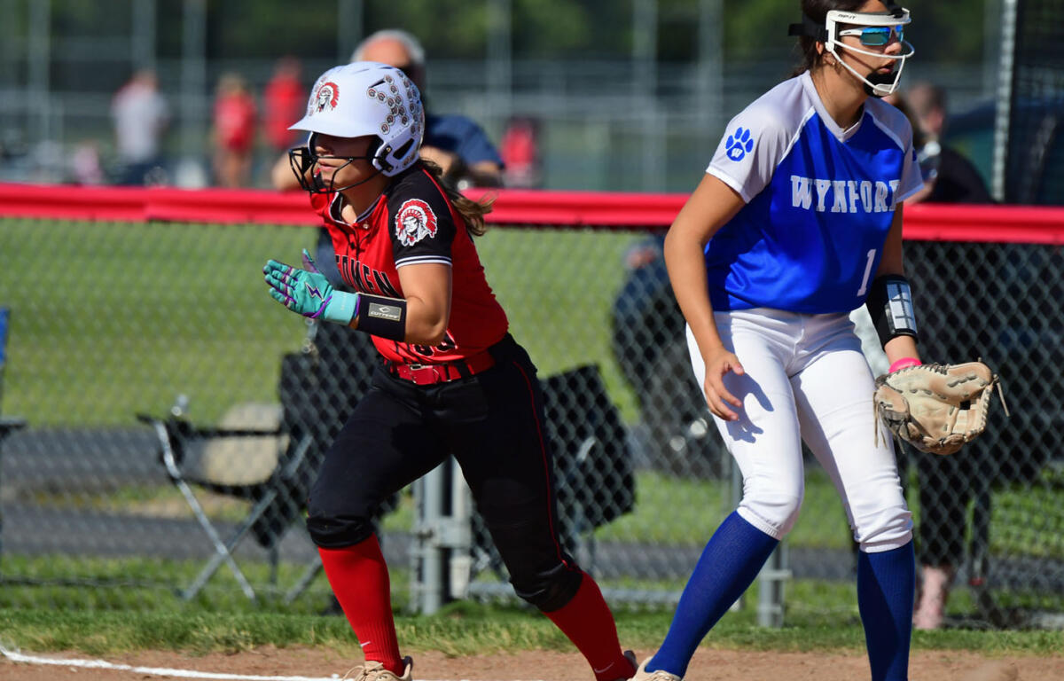 Two female softball players sprint along the basepath: one in red and black with a white helmet, the other in blue and white with a glove.
