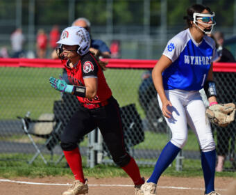 Two female softball players sprint along the basepath: one in red and black with a white helmet, the other in blue and white with a glove.