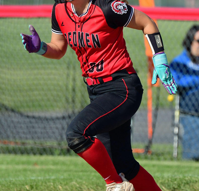 Female softball player sprinting on a dirt field in a red-and-black uniform, white helmet, teal gloves, number 30.