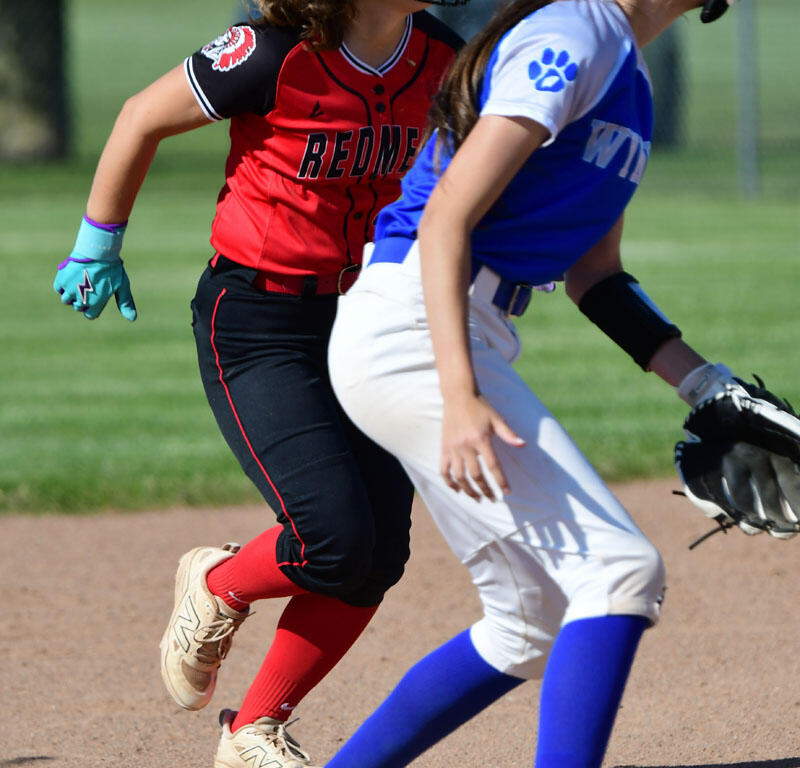 Two young softball players race on a dirt infield, one in a red jersey and helmet behind a blue-and-white runner with a glove ready at first base.