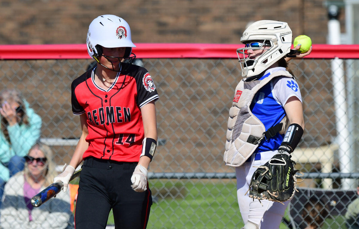 Two softball players on a field: a Redmen batter in red and black with helmet, holding a bat beside a catcher in blue and gray protective gear with a glove and helmet.
