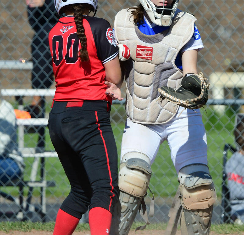 Two girls at a softball game: a catcher in white gear facing a baserunner in red and black near home plate with dust rising from the dirt backstop in the background.