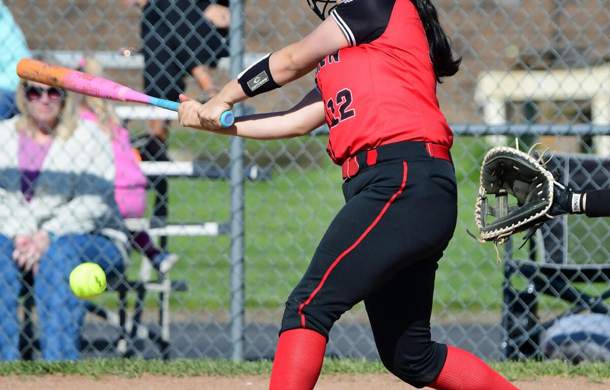 Female softball batter in red and black swings at a pitch during a game, helmet on, glove nearby behind her at home plate.
