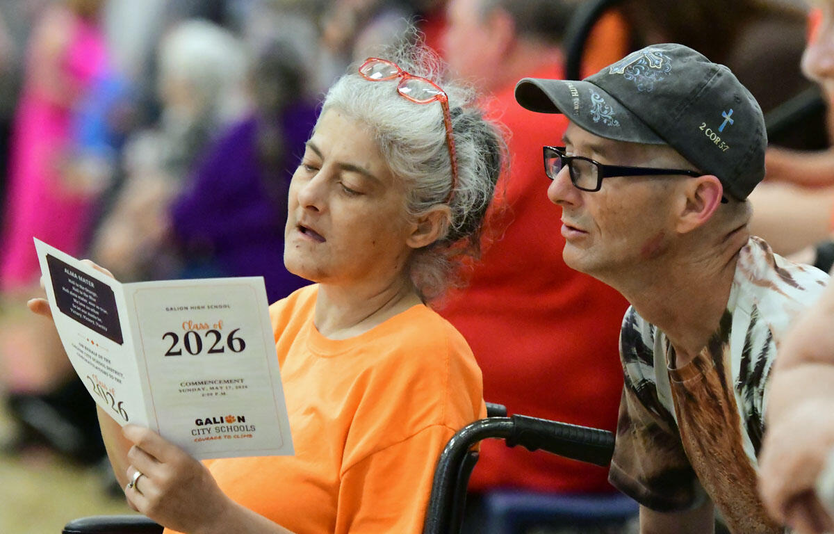 Older woman in an orange shirt reading a graduation program, seated beside a man in a cap at an outdoor event.
