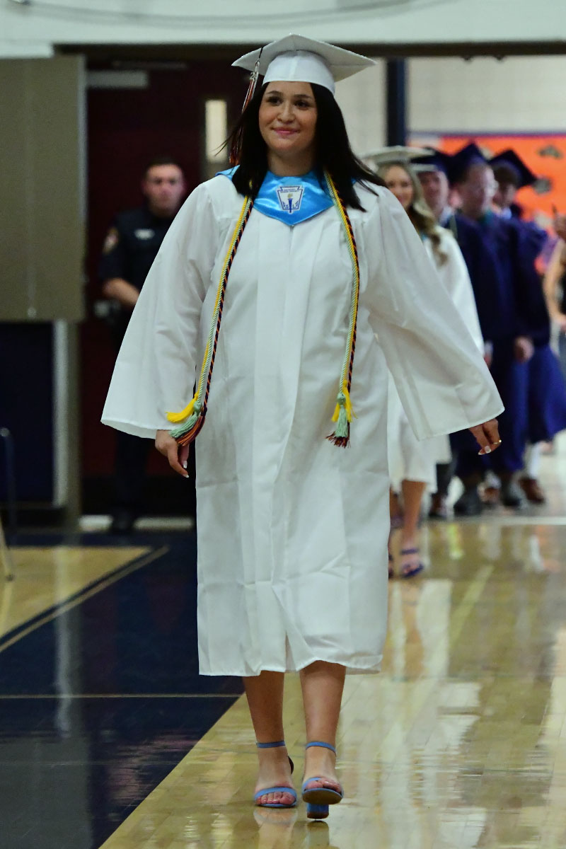 Female graduate in a white cap and gown walking down a hallway, smiling, with honor cords draped over her shoulders