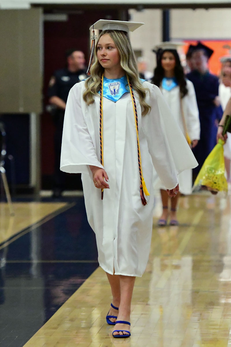 Female graduate in a white gown and cap walking down a gym floor during a commencement ceremony, wearing blue sandals and honor cords.