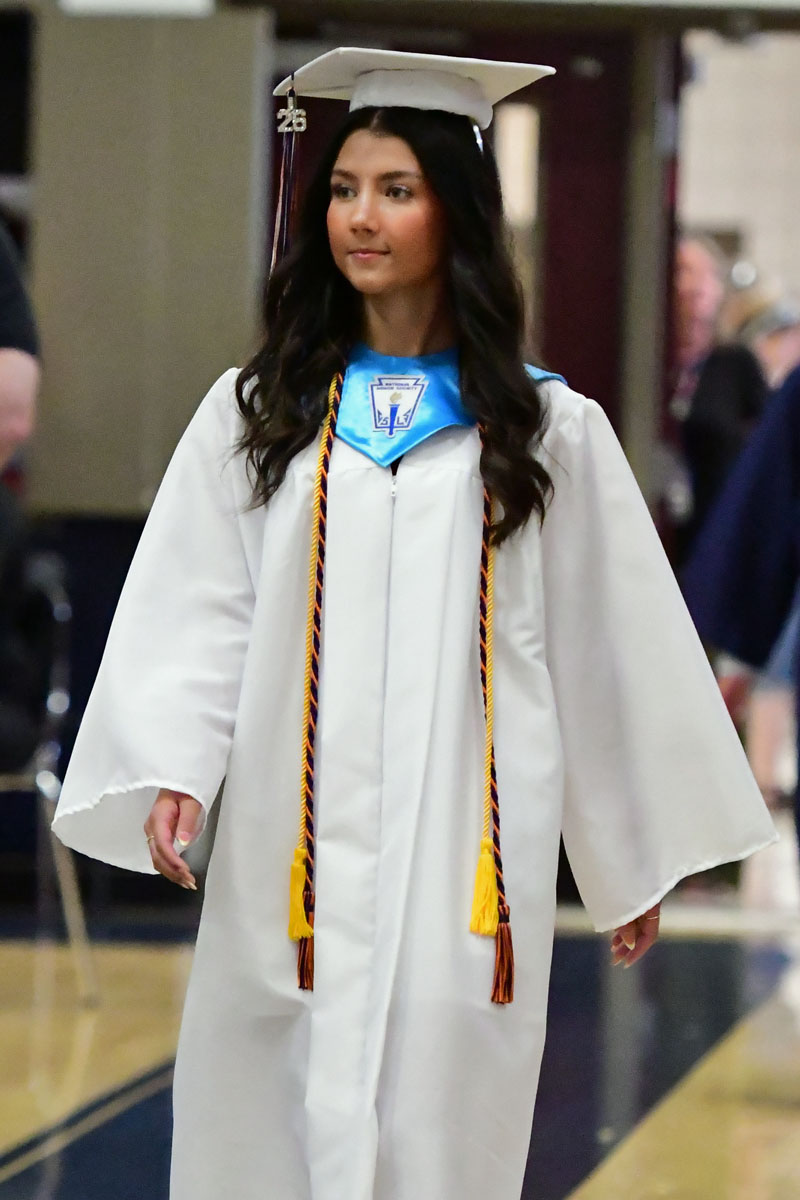 Graduate in white cap and gown walking at a ceremony, with blue honor stole and yellow cords.