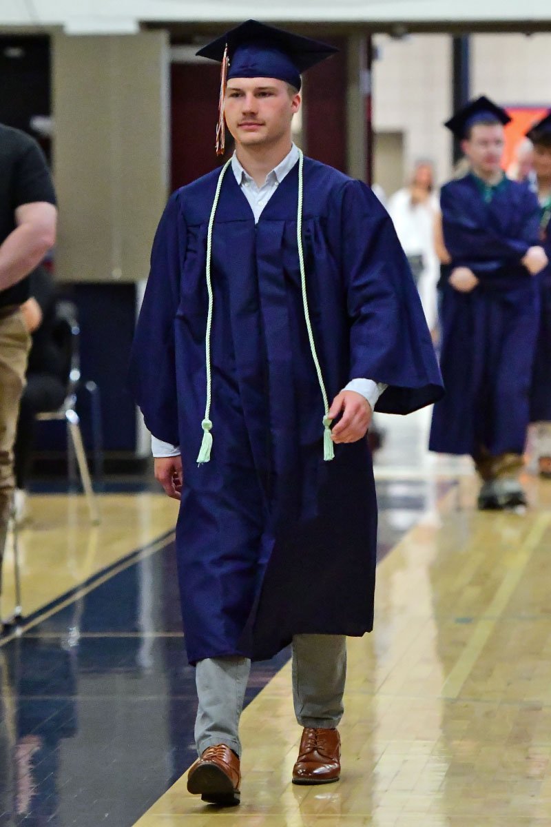 Male graduate in a navy cap and gown walking down a gymnasium aisle, wearing a white shirt and light cords, brown dress shoes visible at the bottom of the gown.