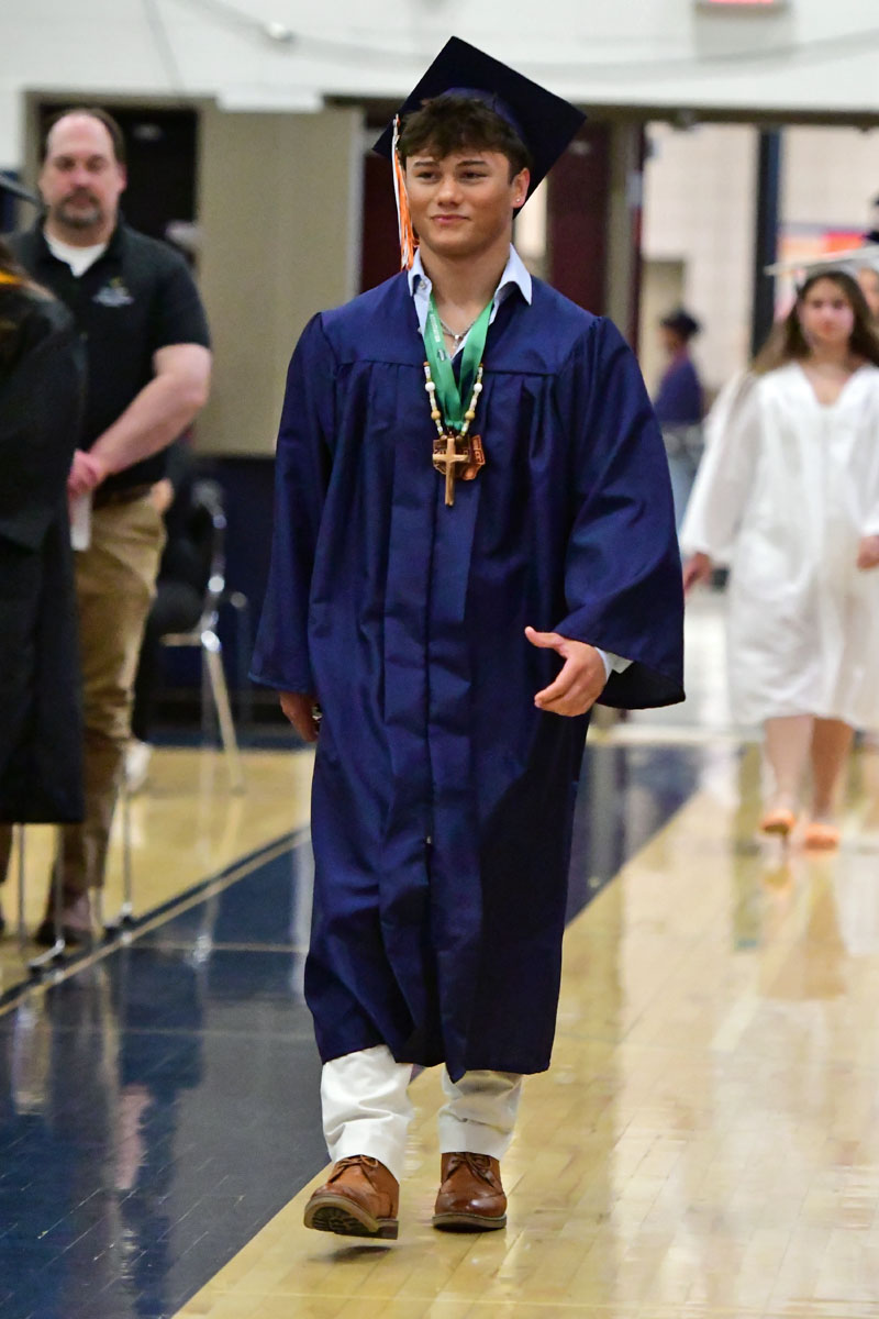 A graduate in a blue cap and gown walking down a gymnasium aisle, wearing a medal and green ribbon.