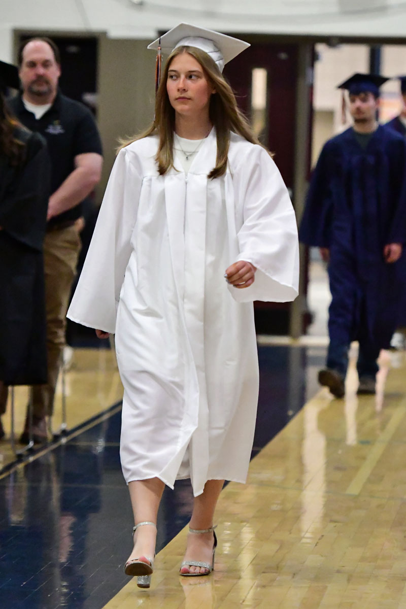 Young graduate in a white gown and cap walking down a hallway at a graduation ceremony.