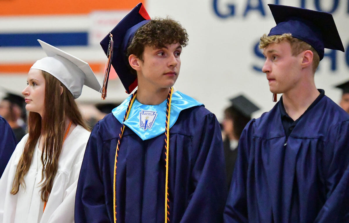 Three graduates in caps and gowns awaiting ceremony: a woman in white on the left, and two men in blue gowns center and right, in a gymnasium setting.