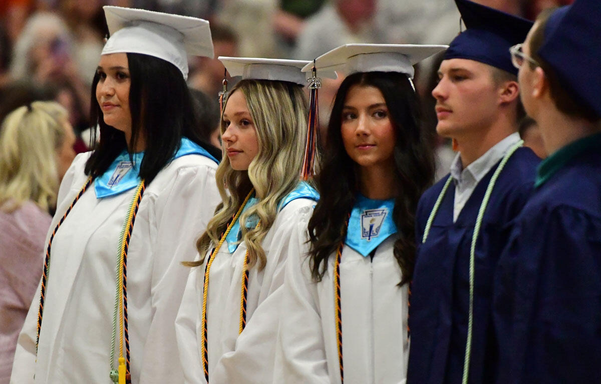 Group of graduates in white gowns and mortarboards standing in line during a ceremony.