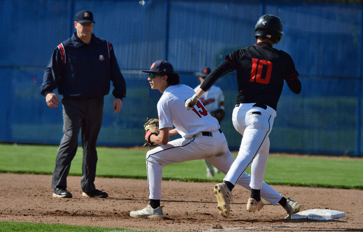 Baseball scene: a fielder in white with number 15 crouches near first base while a runner in black with number 10 slides past the bag; an umpire watches in the background.