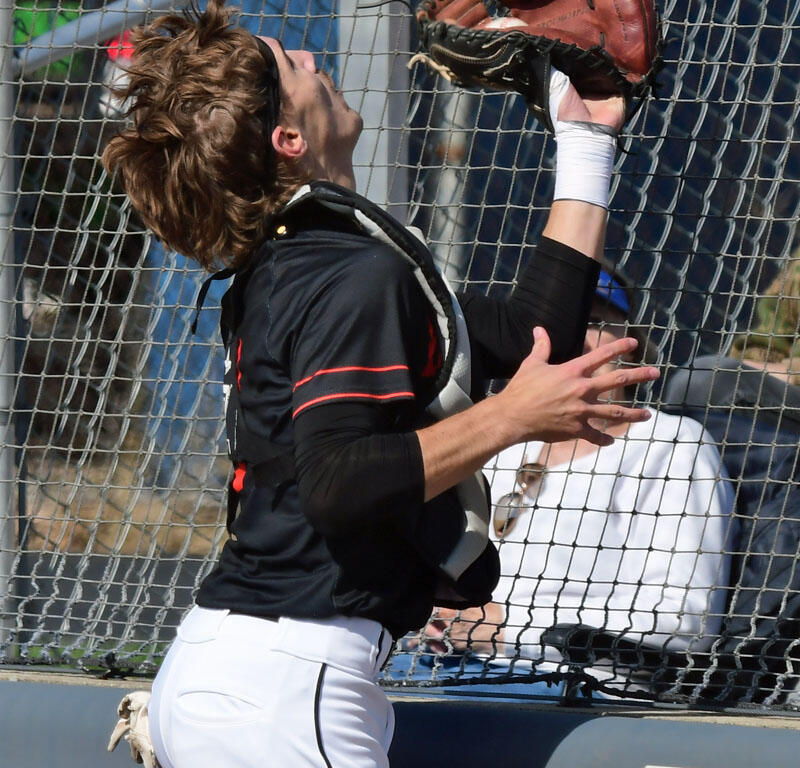 Baseball player in black and white uniform reaches up with a glove to catch a ball near the chain-link fence.