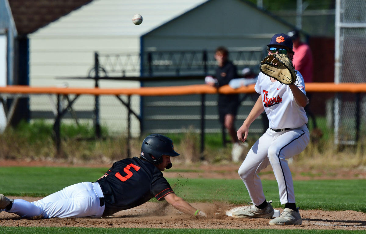 Baseball: a sliding runner in black jersey with orange number 5 dives head-first toward a base as a fielder in white with orange lettering and a tiger paw glove prepares to catch an incoming ball nearby.