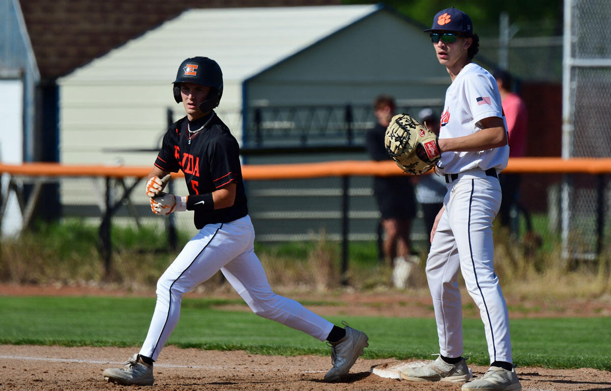 A baseball baserunner in a black jersey reaches first base while the fielder waits nearby with a glove on the base.”,
