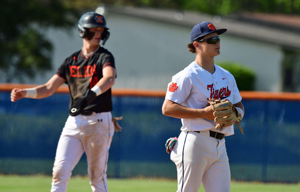 Two baseball players on a field during a game: a Clemson Tigers player in white with a glove in the foreground and a rival in a black jersey in the background.