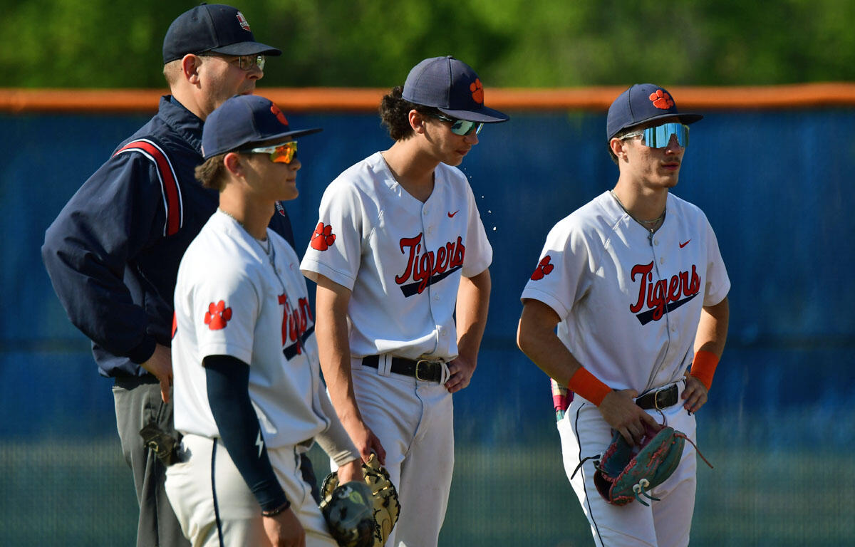 Baseball players in white Tigers uniforms with orange paw prints stand with gloves on a sunny field, coach in background.