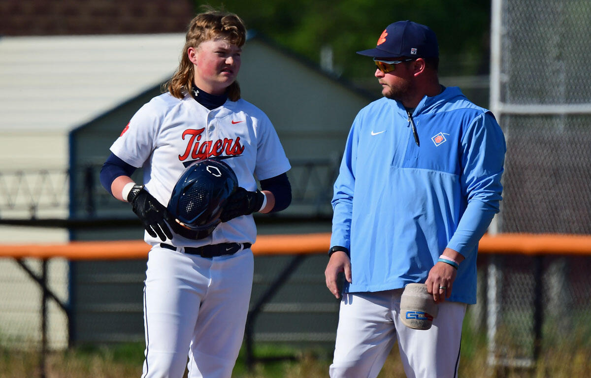 Baseball player in a white Tigers uniform holds a helmet while talking with a coach in a blue jacket on a sunny field.