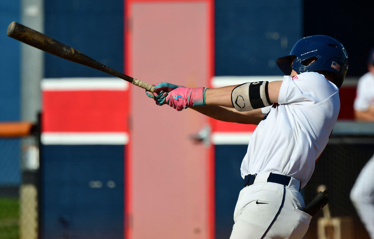 Baseball batter in white uniform and blue helmet mid-swing with a wooden bat, pink gloves visible.