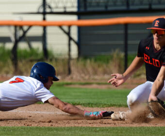 Baseball play action: a white-uniformed runner slides headfirst toward a base as a fielder in black reaches with a glove; dirt kicks up around them.