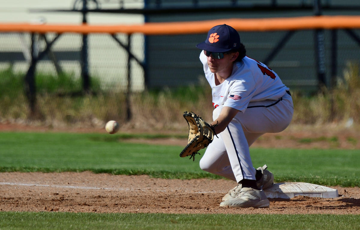 Baseball fielder in a white uniform and navy cap with an orange paw print stretches to field a ground ball near first base.