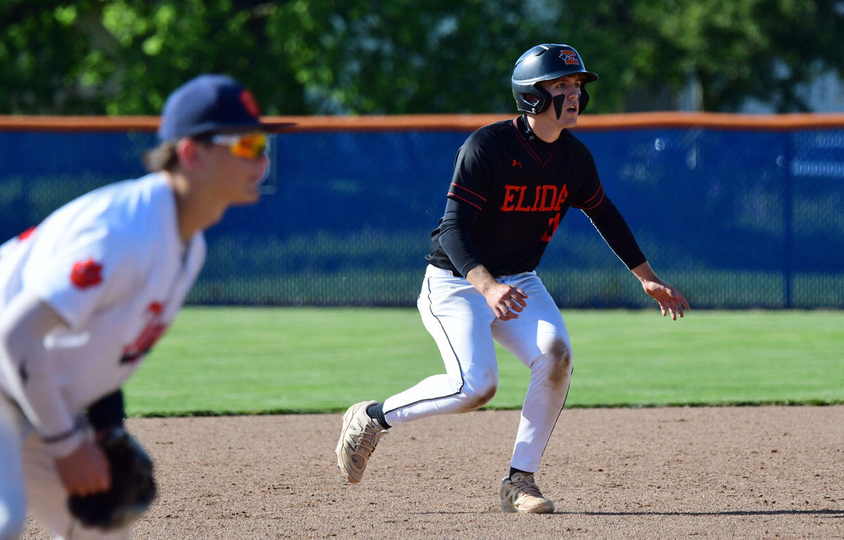 Baseball player in black ELIDA uniform runs on the infield as a fielder in white tries to tag from the left, during a game on a sunny day.