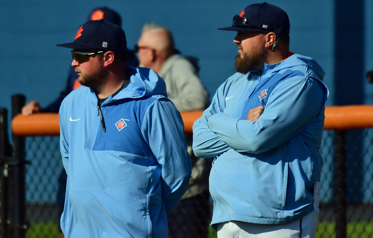 Two men in light-blue jackets and dark caps stand side by side at a baseball field, arms crossed, watching a game.