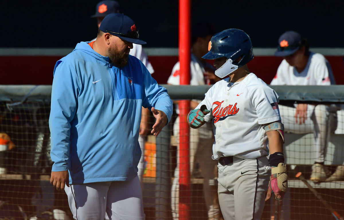 Baseball coach in a blue jacket high-fives a player in a white uniform near the dugout railing.