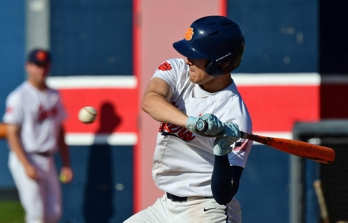 Baseball batter in white uniform swings at a pitched ball, helmet on, orange bat ready to hit on a field with red and blue background.