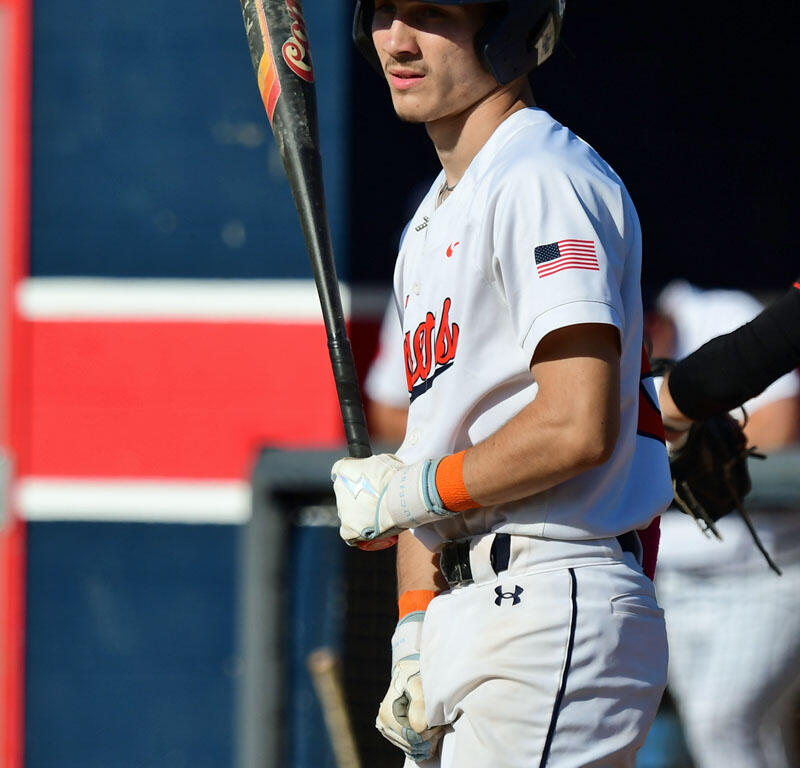 Baseball player in white uniform with an orange paw print helmet logo, gripping a bat at the dugout entrance.
