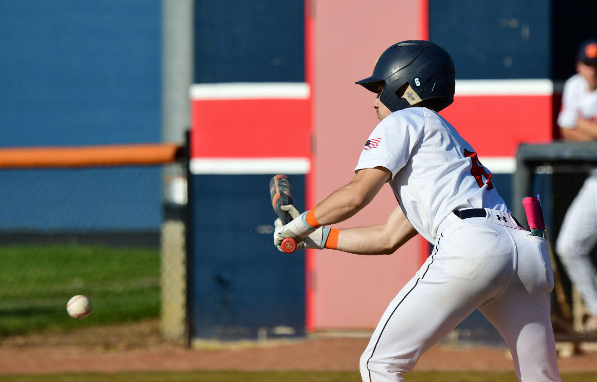 Baseball player in a white uniform and helmet swings at a pitch, with a ball in midair nearby on a sunny field.