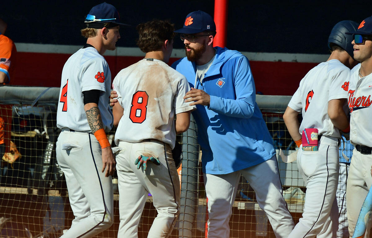Baseball players in white uniforms stand near the dugout as a bearded coach in a blue jacket talks to a player wearing #8 with a dirt-stained back.