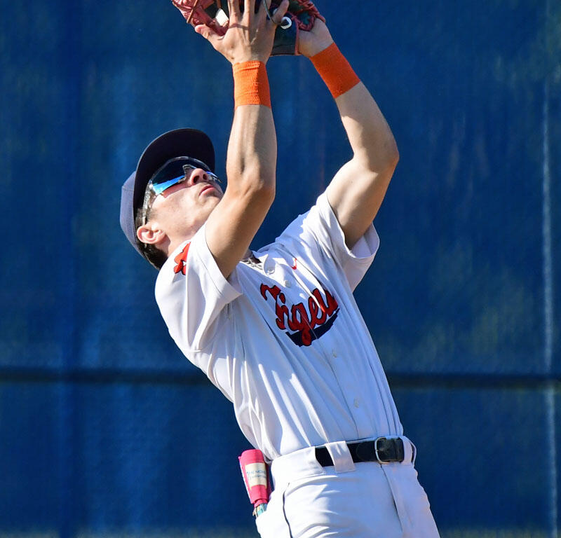 Baseball player in white uniform reaching up to catch a ball with a glove, outdoors on a blue fence background.