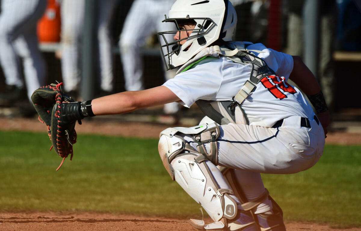 Baseball catcher in white protective gear crouches behind home plate, glove extended to catch the pitch on the dirt infield.