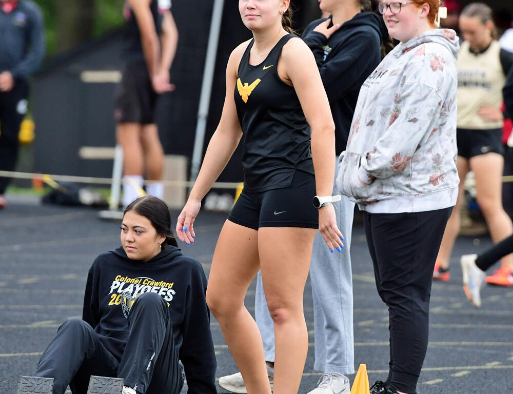 Athletes waiting at a track starting area: a woman in black athletic wear stands while another sits on the track near starting blocks and cones.