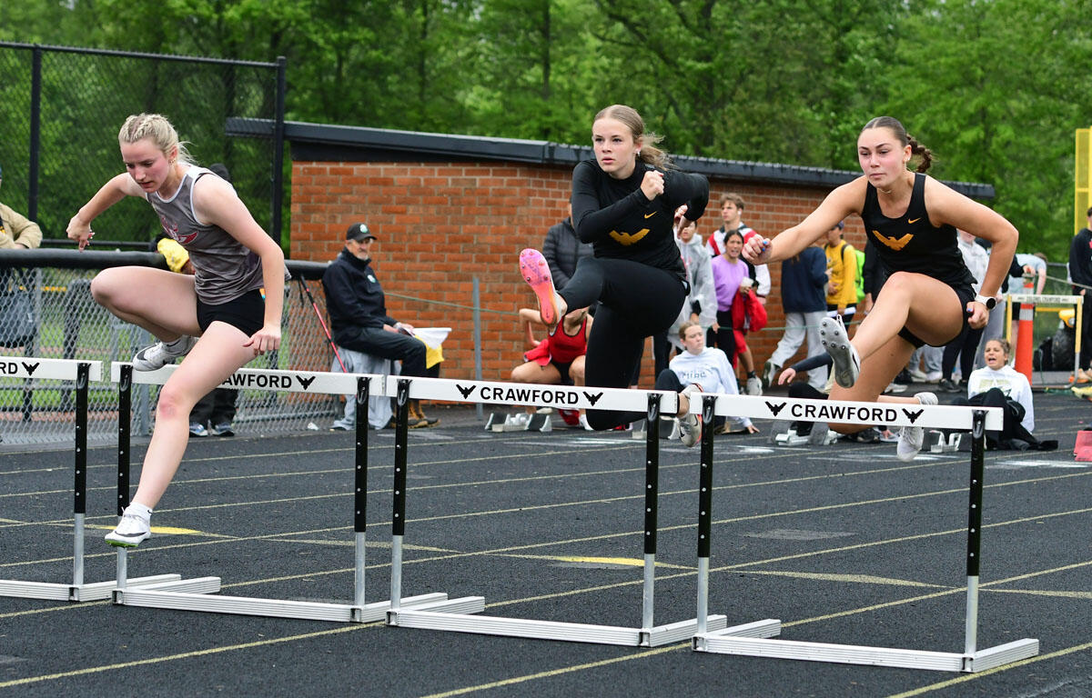 Three female hurdlers leap over hurdles on a track during a race, with spectators and a brick wall in the background.