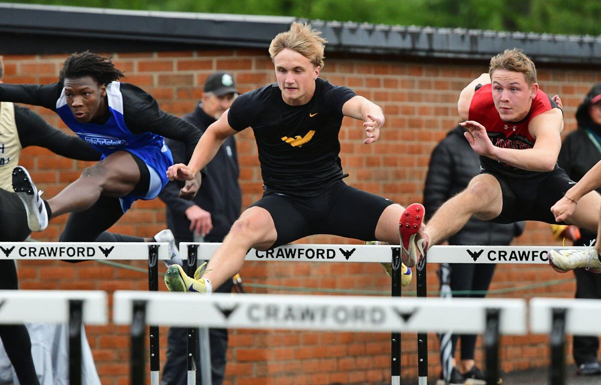 Three teenage hurdlers clear hurdles in a race, mid-air over a white Crawford barrier against a brick wall backdrop.